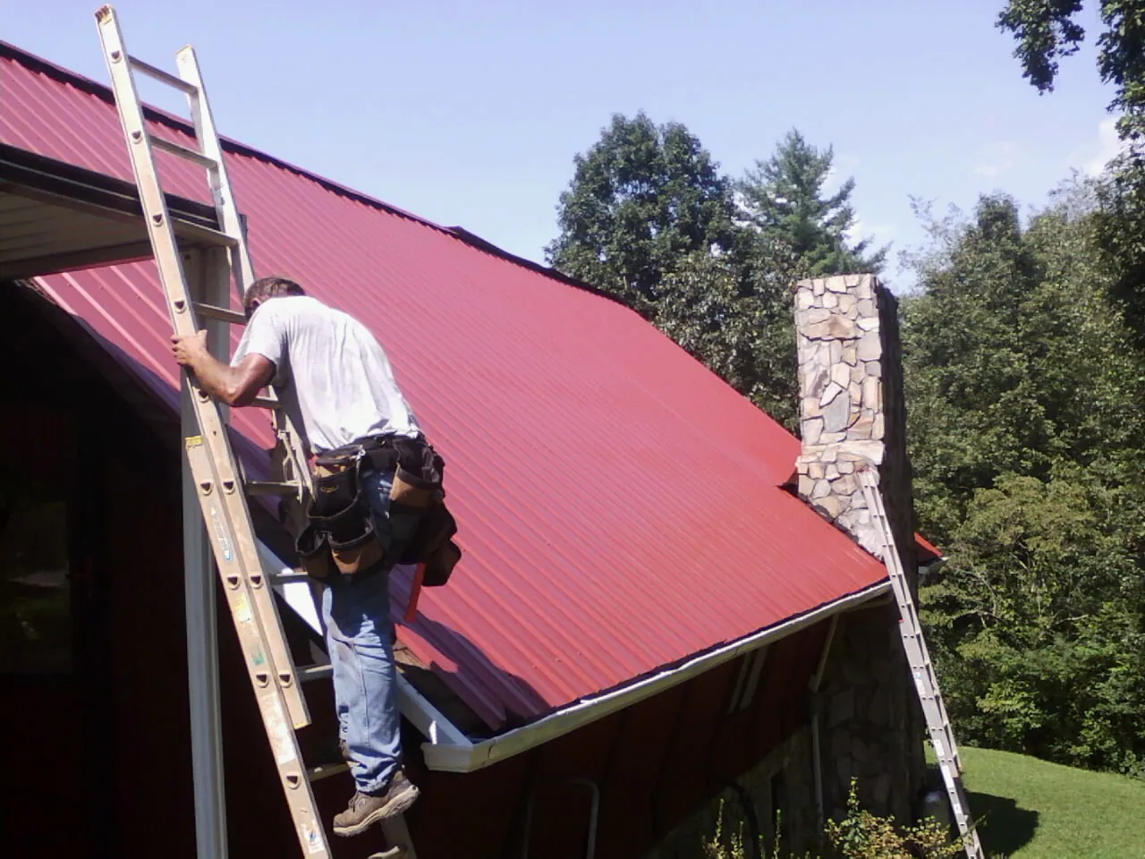 Roofer on a ladder beside a red metal roof and stone chimney — Soffit & Fascia Repair by Roofing Brothers in Niantic